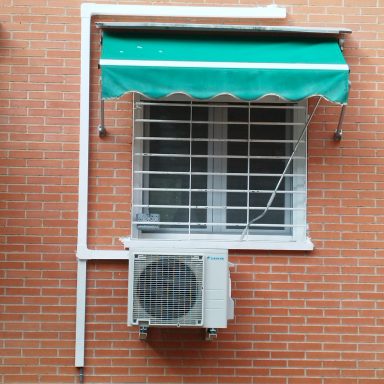 Ventana con reja, toldo verde y aire acondicionado en pared de ladrillo.